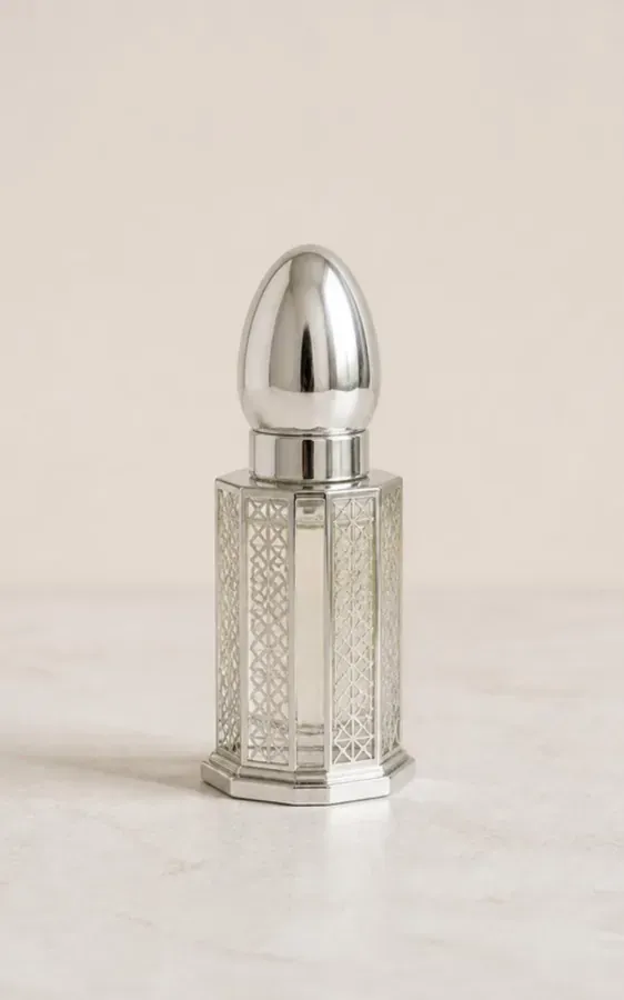 Petit flacon d’huile parfumée en verre transparent à facettes orné de bandes argentées, bouchon argenté en forme d’œuf, posé seul sur une pierre claire